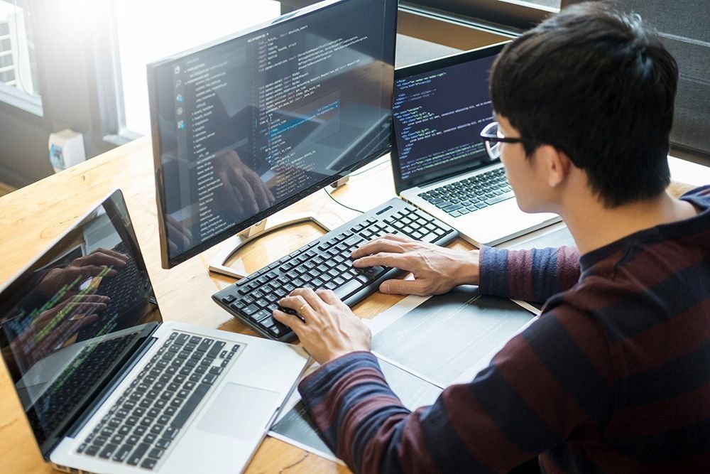 Ein Mann sitzt vor 3 Bildschirmen, auf denen ein Code zu sehen ist | CDA IT Systems GmbH